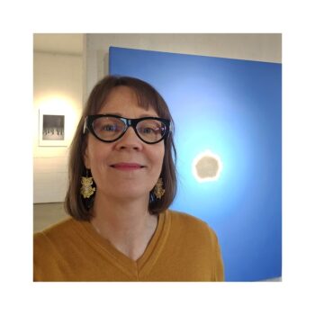 A caucasian woman stands infront of an oil painting in a gallery space. She's the artist Outi Kotala and the painting is made by her. It depicts a small cloud that infront of the sun on a wide blue sky.